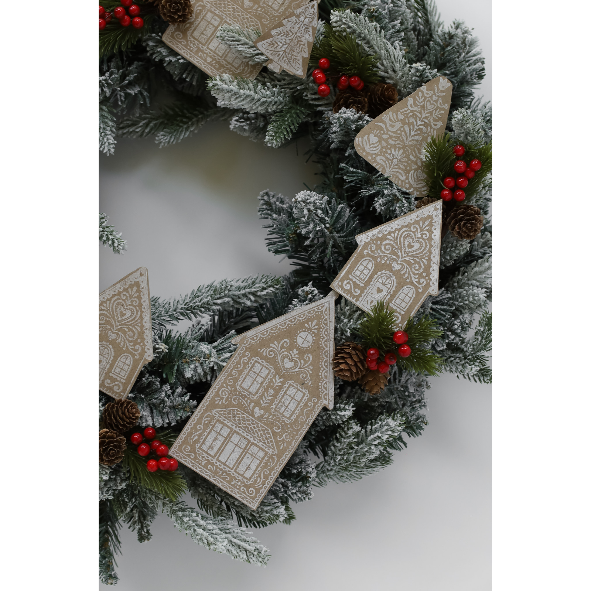 A festive Christmas wreath with frosted green pine branches, red berries, pinecones, and hanging gingerbread house ornaments on a light gray background.