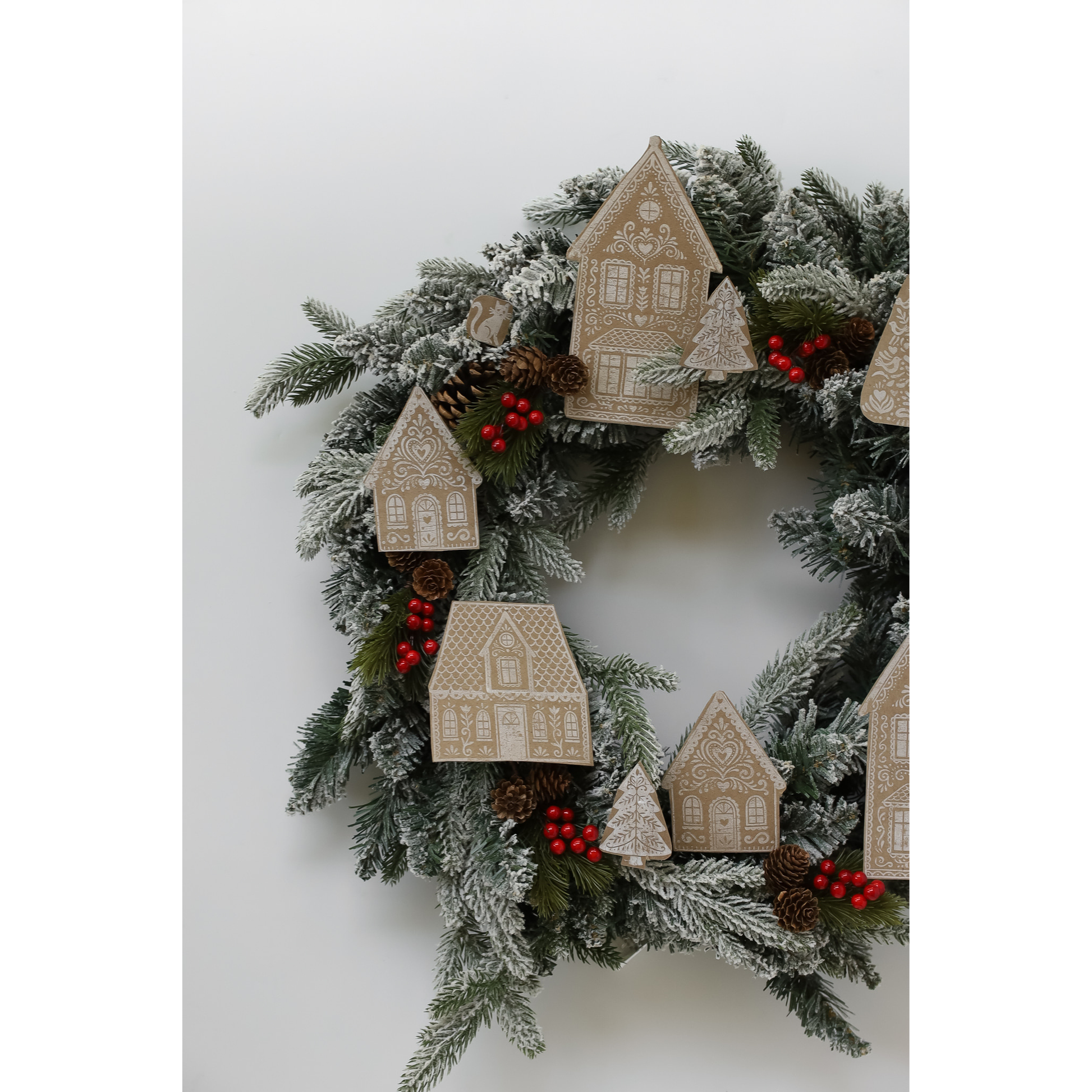 A festive Christmas wreath with frosted green pine branches, red berry clusters, pinecones, and beige wooden house ornaments, shown against a plain light gray background.