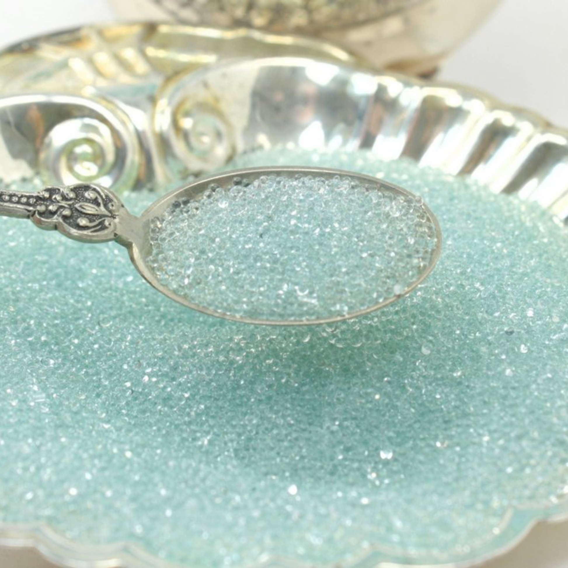 Silver spoon holding small clear glass beads decorative silver rimmed dish in the background.