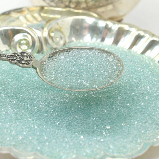 Silver spoon holding small clear glass beads decorative silver rimmed dish in the background.