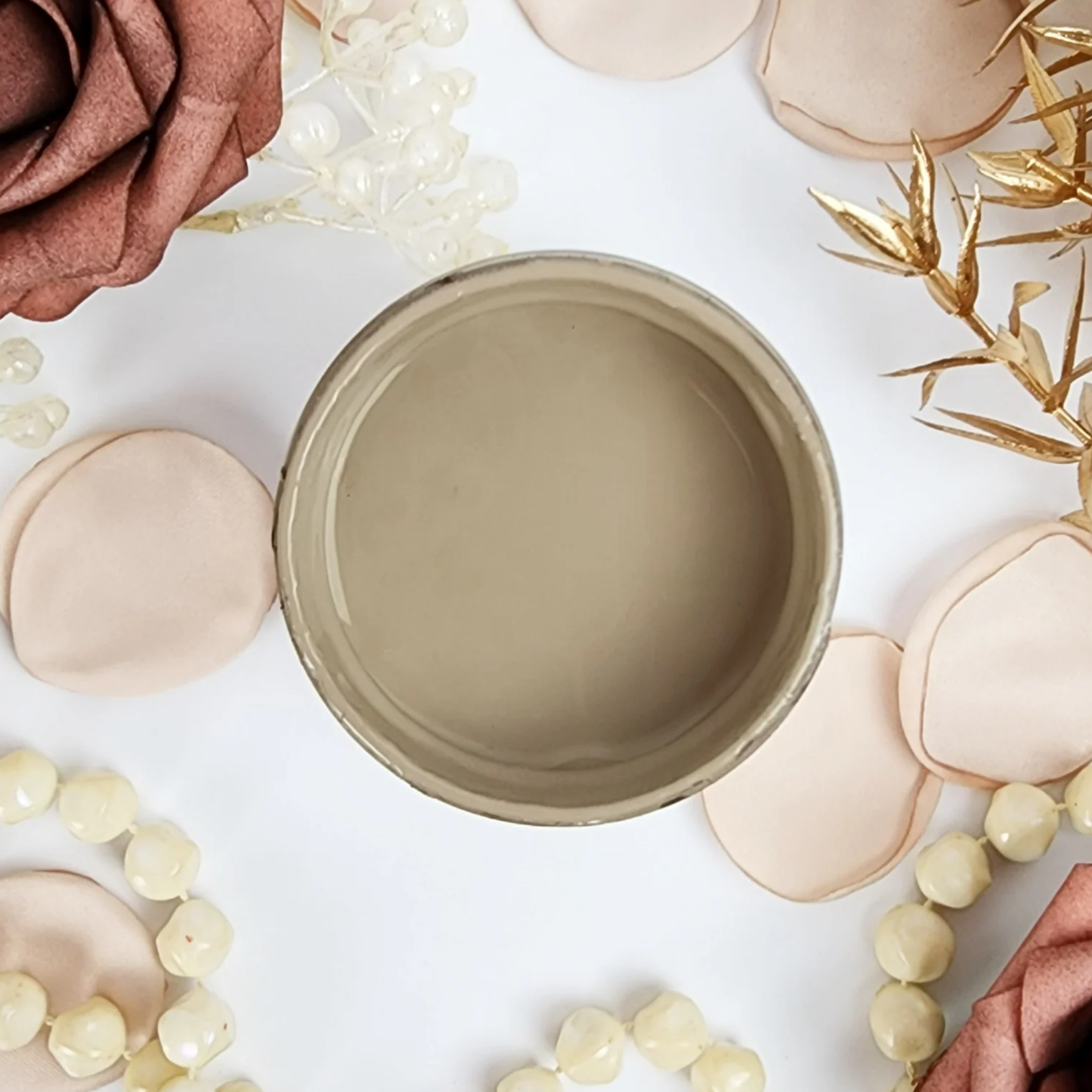 Image of open jar of light brown colored paint with peach rose petals, brown rose and beads on a white background.
