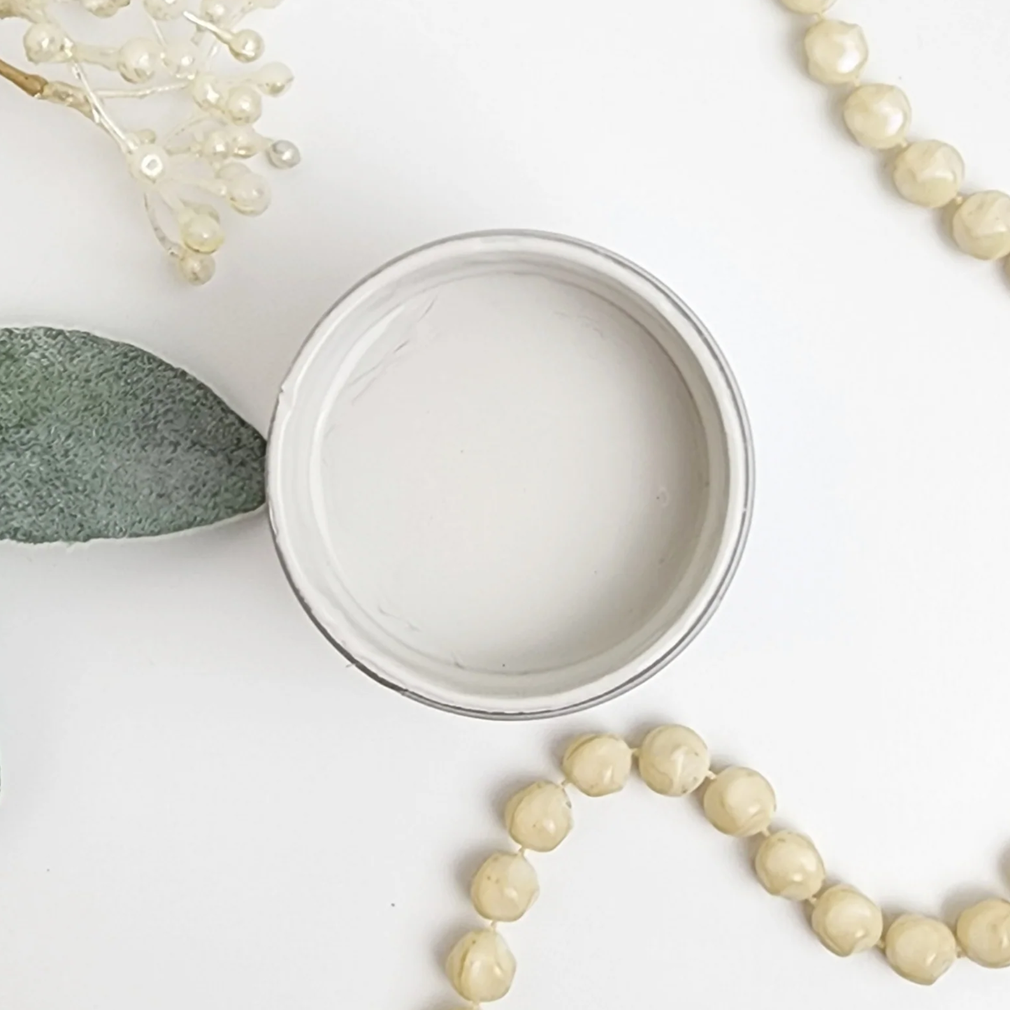 Image of open jar of off white paint with beige beeds and leaves on a white background.