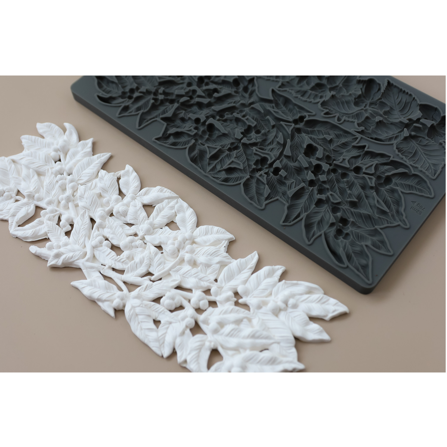 A dark gray rectangular silicone mold with embossed floral leaf patterns, shown next to a long strip of white molded floral leaf trim on a beige background.