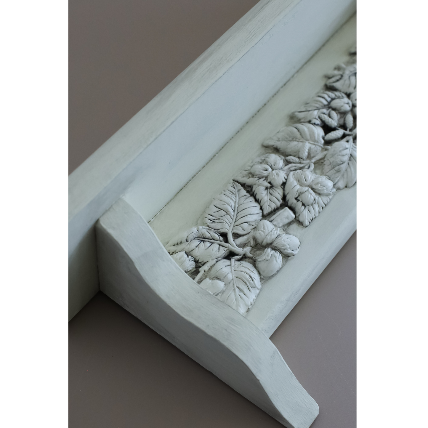 Close-up of a white decorative wall shelf with a raised leaf motif, shown against a plain taupe background.