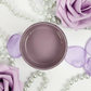 Image of open jar of medium lavender/purple paint with lavender flower petals, lavender roses and string of pearls on a white background.