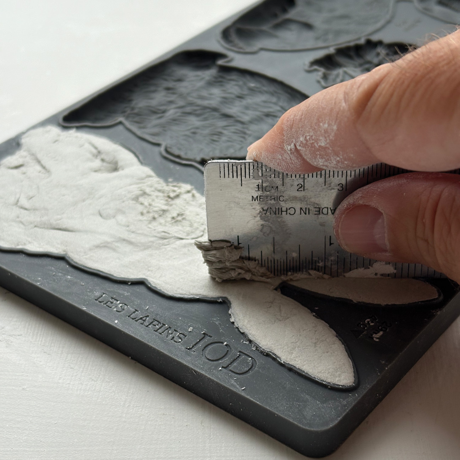 Hand using a ruler to level a piece of white clay on a silicone mold.
