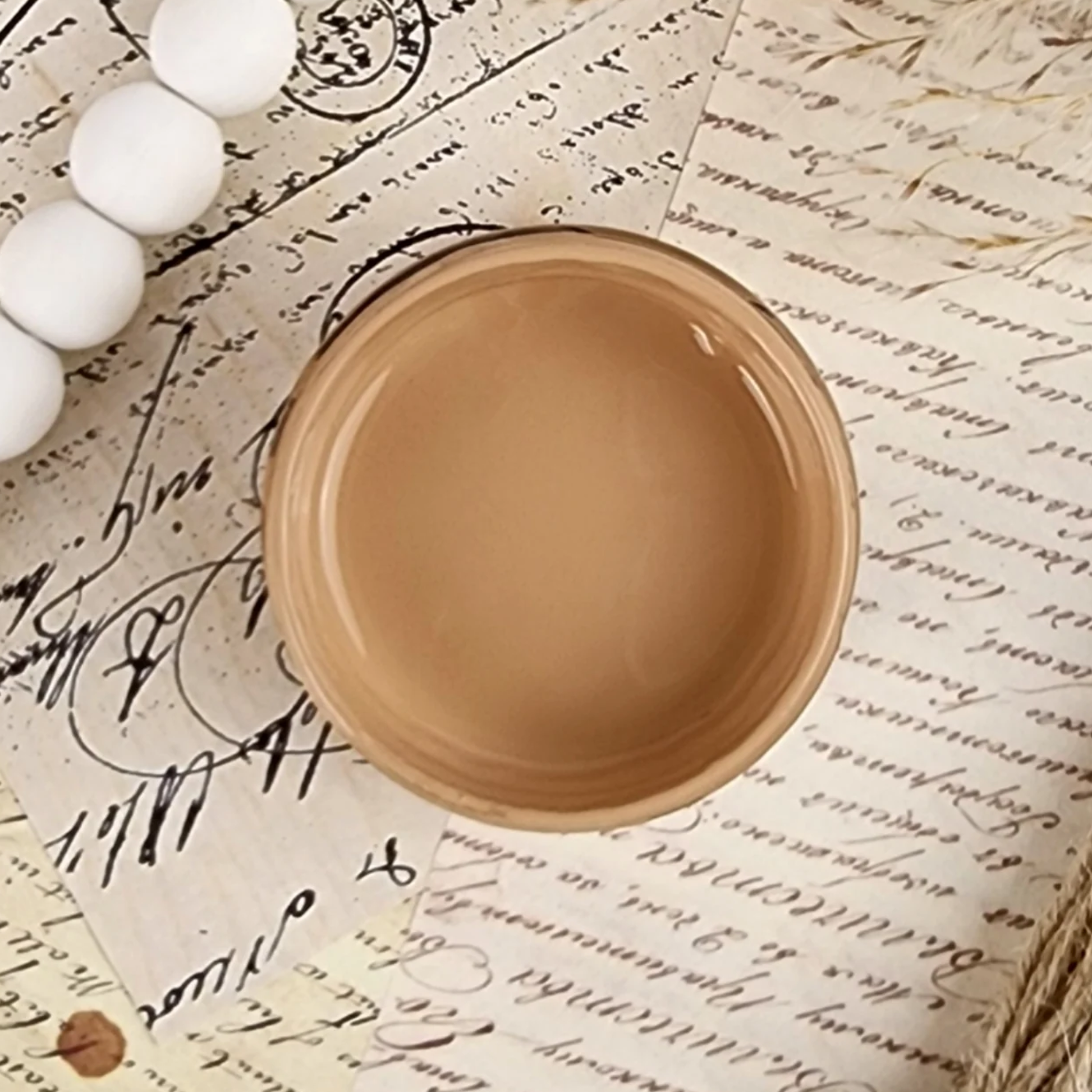 Image of open jar of golden beige colored paint on sepia toned cursive text ephemera.