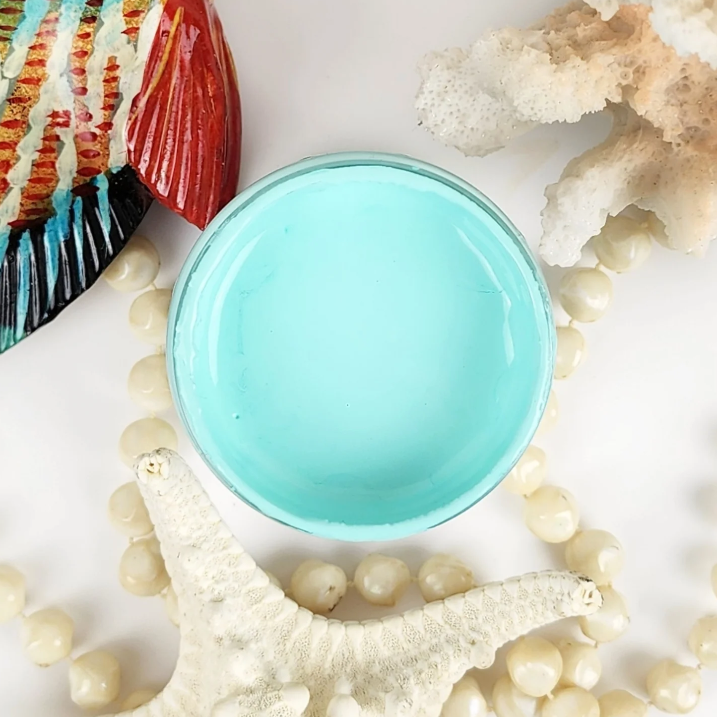 Image of open jar of pale aqua colored paint with sea shells, a colorful ceramic fish and beige pearls on a white background.