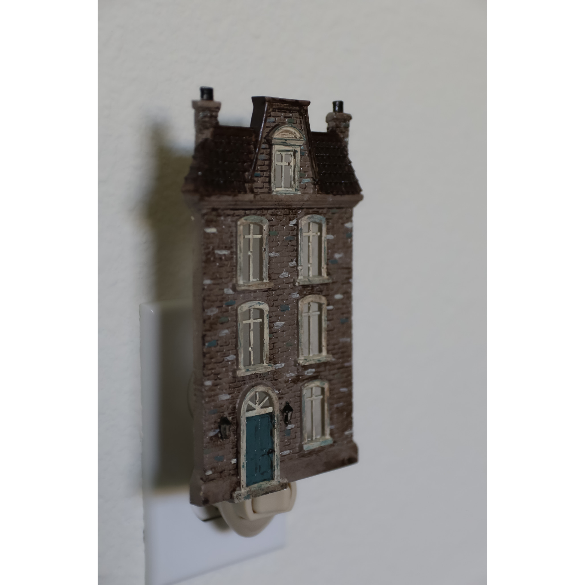A miniature decorative house-shaped night light plug-in, featuring a detailed brown brick facade, white windows, and a blue door, shown mounted on a white wall.