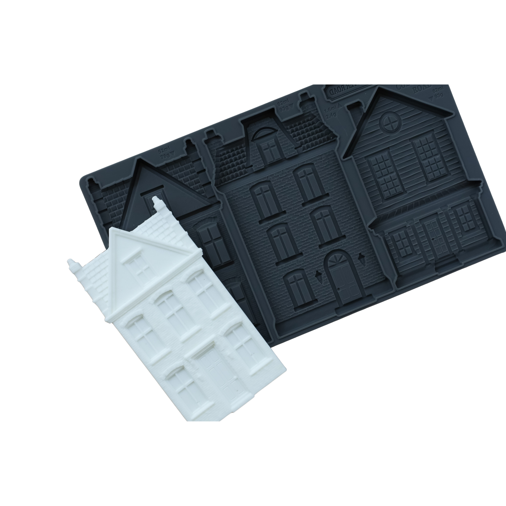 A dark gray silicone mold with detailed house-shaped cavities, shown with a small white house-shaped product sample in front, on a plain white background.