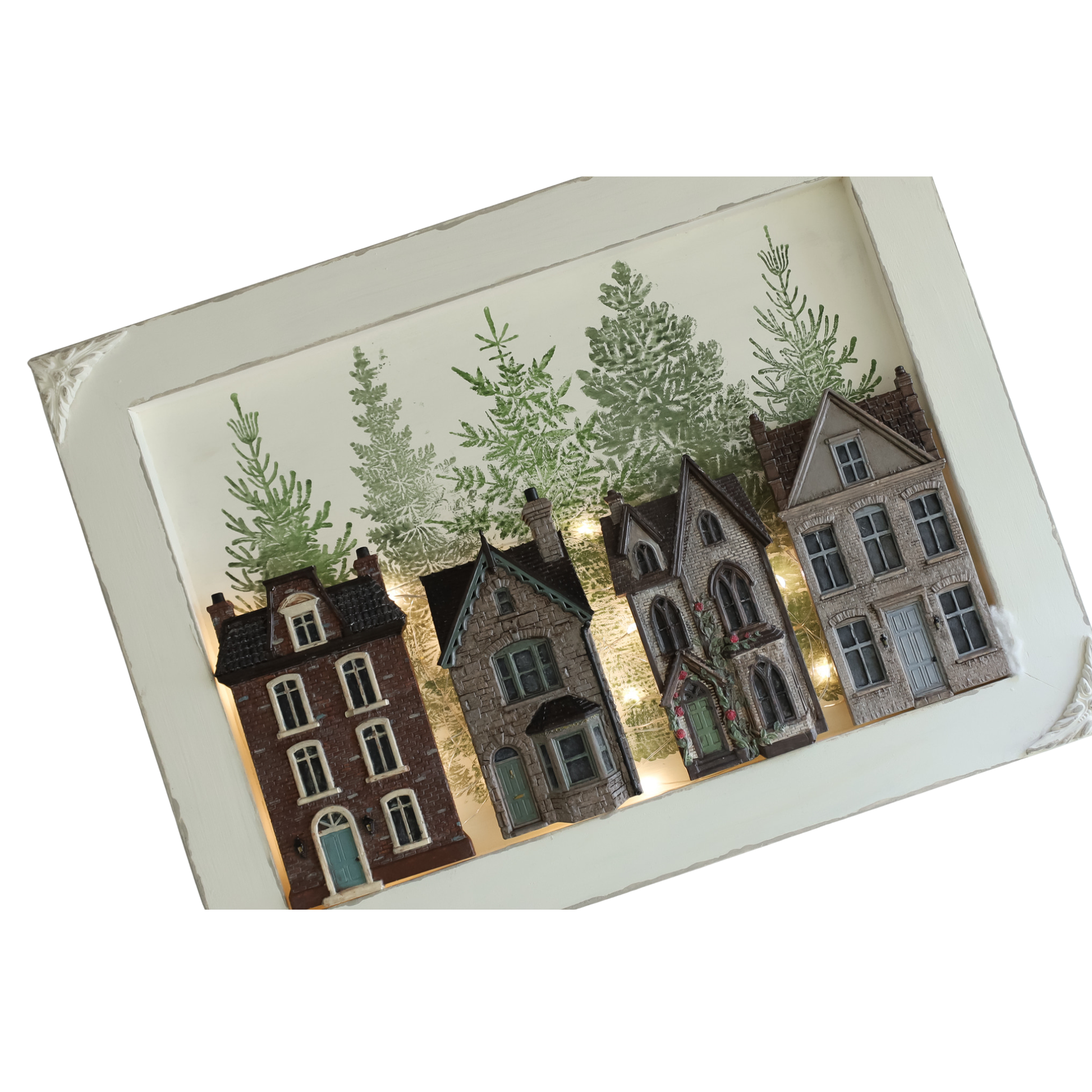 A rectangular decorative wall art piece featuring a row of detailed miniature-style houses in brown, gray, and beige tones, with green trees above and a white border around the artwork.