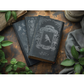 Three darkly colored books with ornate designs on a rustic wooden surface with green leaves.