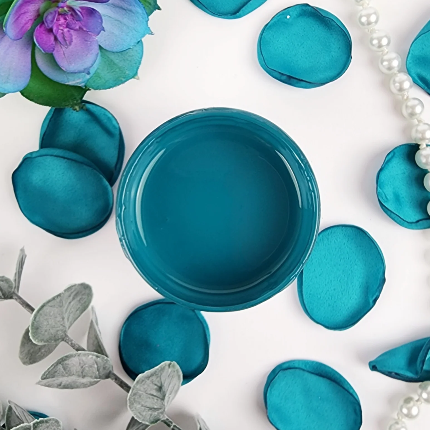 Image of open jar of deep turquoise paint with blue flower petals and pearls.