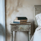Stack of books on a weathered wooden nightstand next to a bed with white bedding.