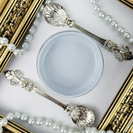 Image of open jar of pale blue paint with pearls, silver spoons and a gold frame in the background.