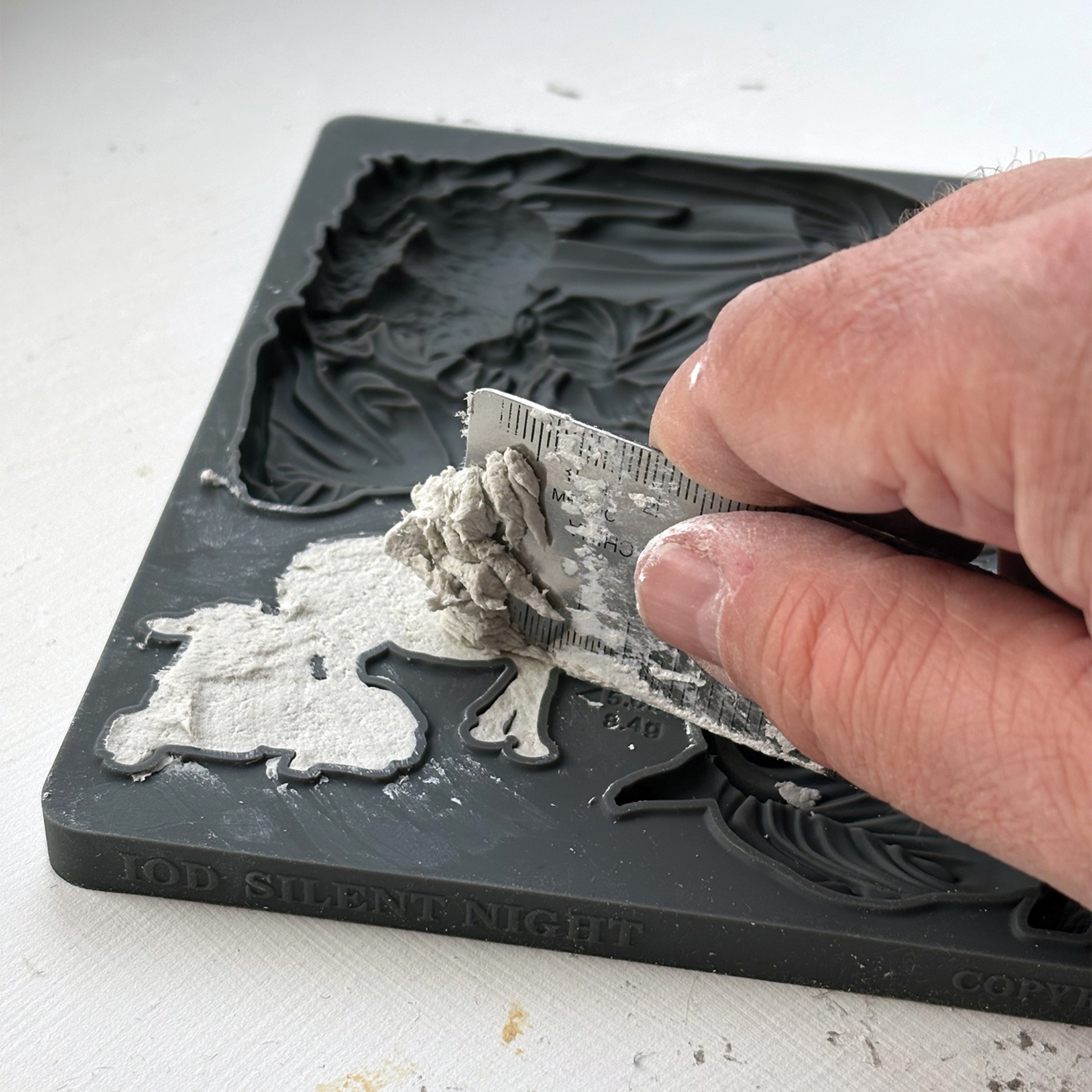 A close-up of a hand using a small metal ruler to press clay into a silicone mold with the embossed text 'IOD SILENT NIGHT' visible on the mold.