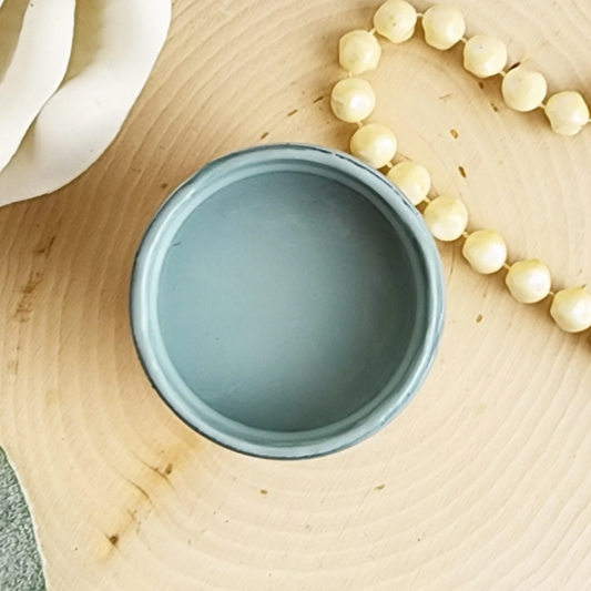 Image of an open jar of pale blue paint on a wood background with a stringed necklace.