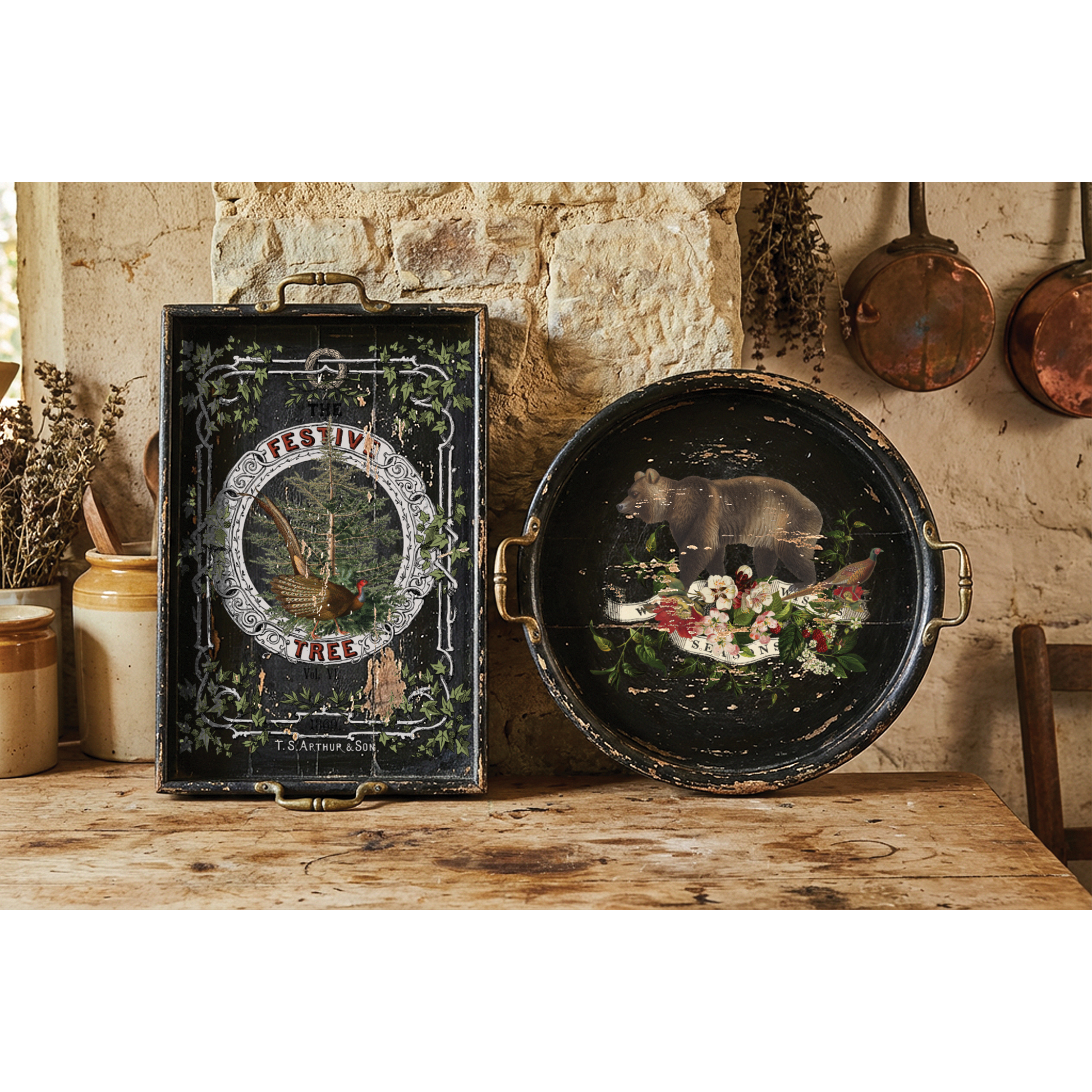 Two decorative vintage-style metal trays displayed on a rustic wooden table against a stone wall. One rectangular tray features a black background with ornate green vine borders and a central circular illustration reading 'FESTIVE TREE.'
