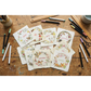 A flat-lay arrangement of twelve square cream paper cards with rustic edges, each printed with a different month's floral wreath and small animal illustrations, displayed on a weathered wooden tabletop surrounded by art supplies.