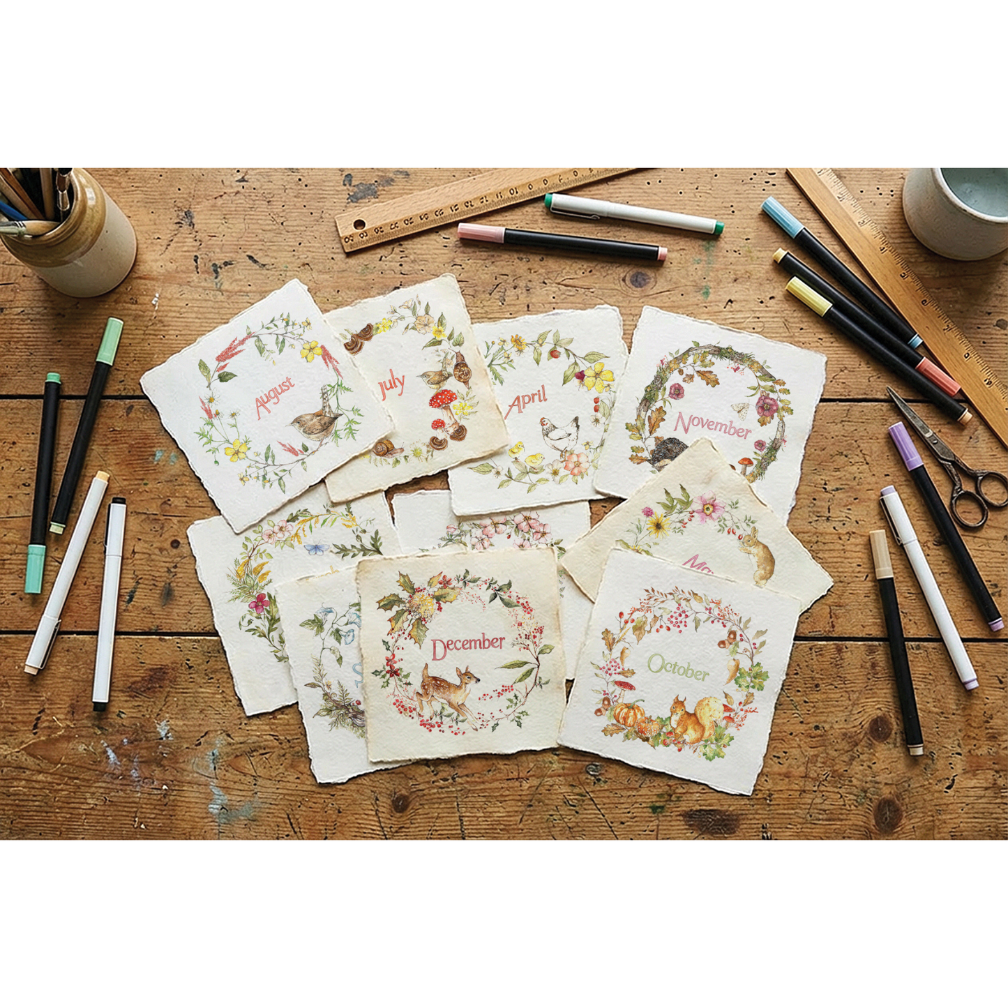 A flat-lay arrangement of twelve square cream paper cards with rustic edges, each printed with a different month's floral wreath and small animal illustrations, displayed on a weathered wooden tabletop surrounded by art supplies.