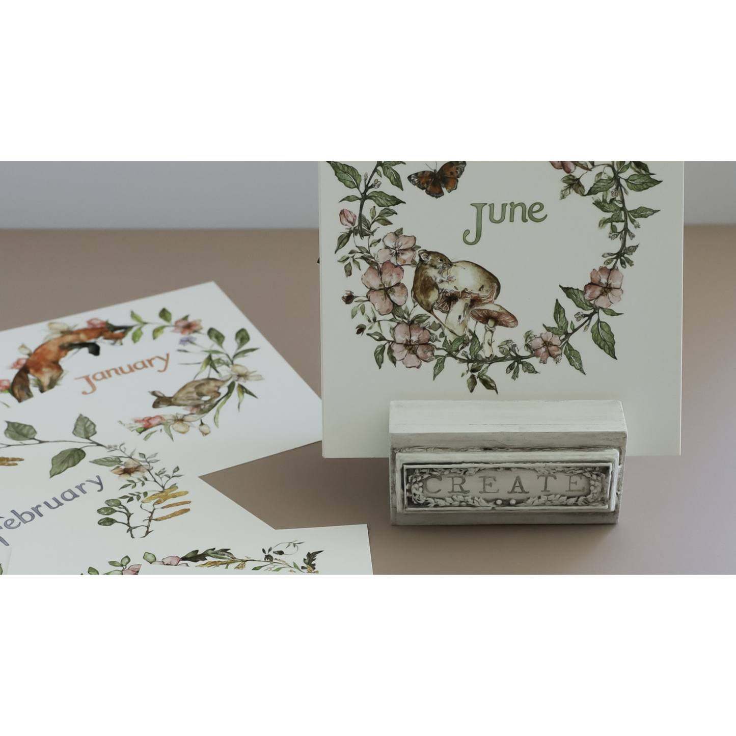 A set of imonthly calendar cards displayed on a small stand, with one card standing upright showing June and others partially visible showing January and February. The cards feature vintage botanical and animal IOD transfers in muted green, pink, and brown tones on an off-white background.