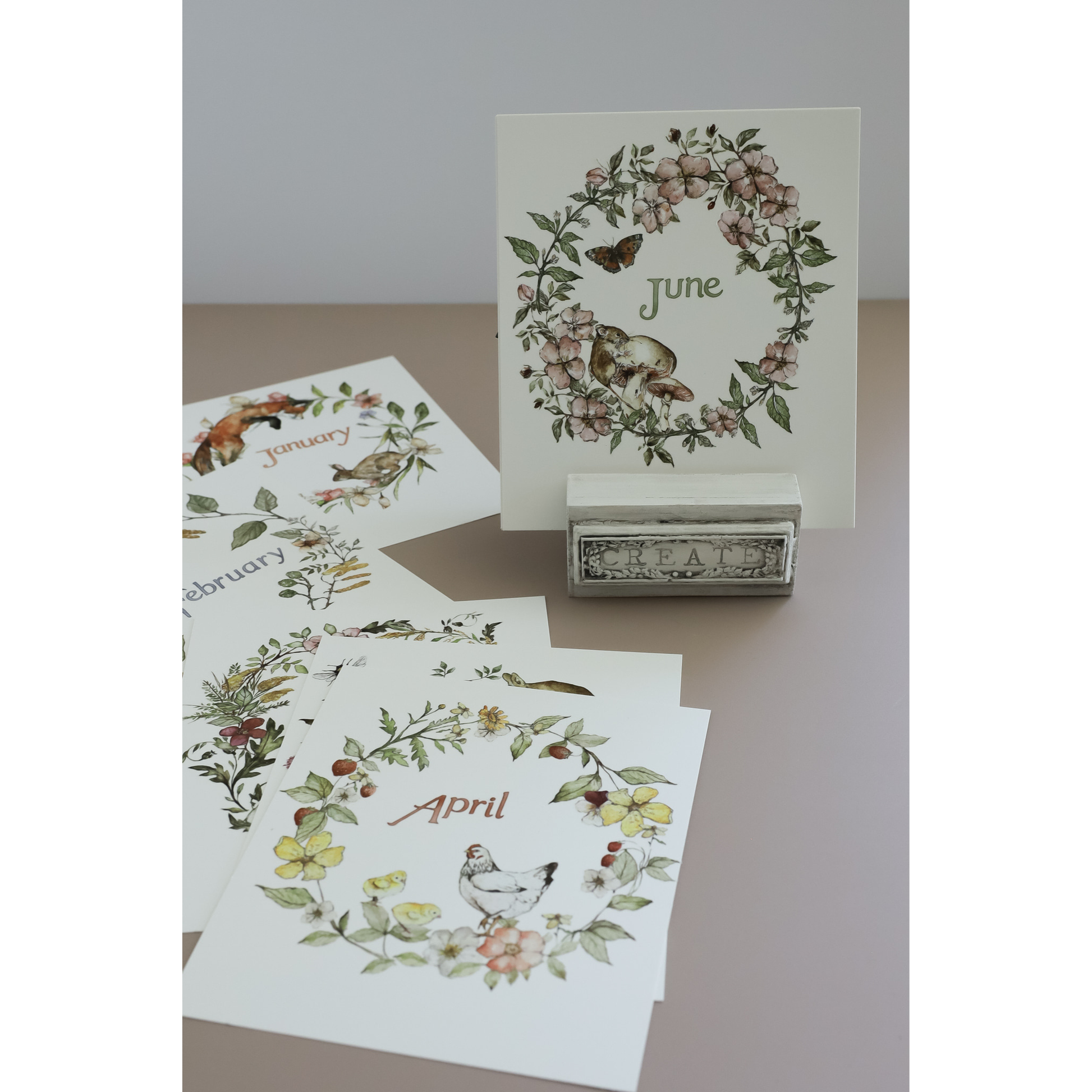 A set of monthly cards displayed on a tabletop, with one card standing upright on a small stand and several cards fanned out below. The cards feature watercolor floral wreaths and seasonal IOD transfer images, including a chicken for April and a fox for January.
