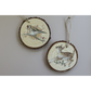 Two round wooden slice ornaments hanging on a light gray wall. One ornament features a goldfinch perched on a branch, and the other features a brown-and-white deer.