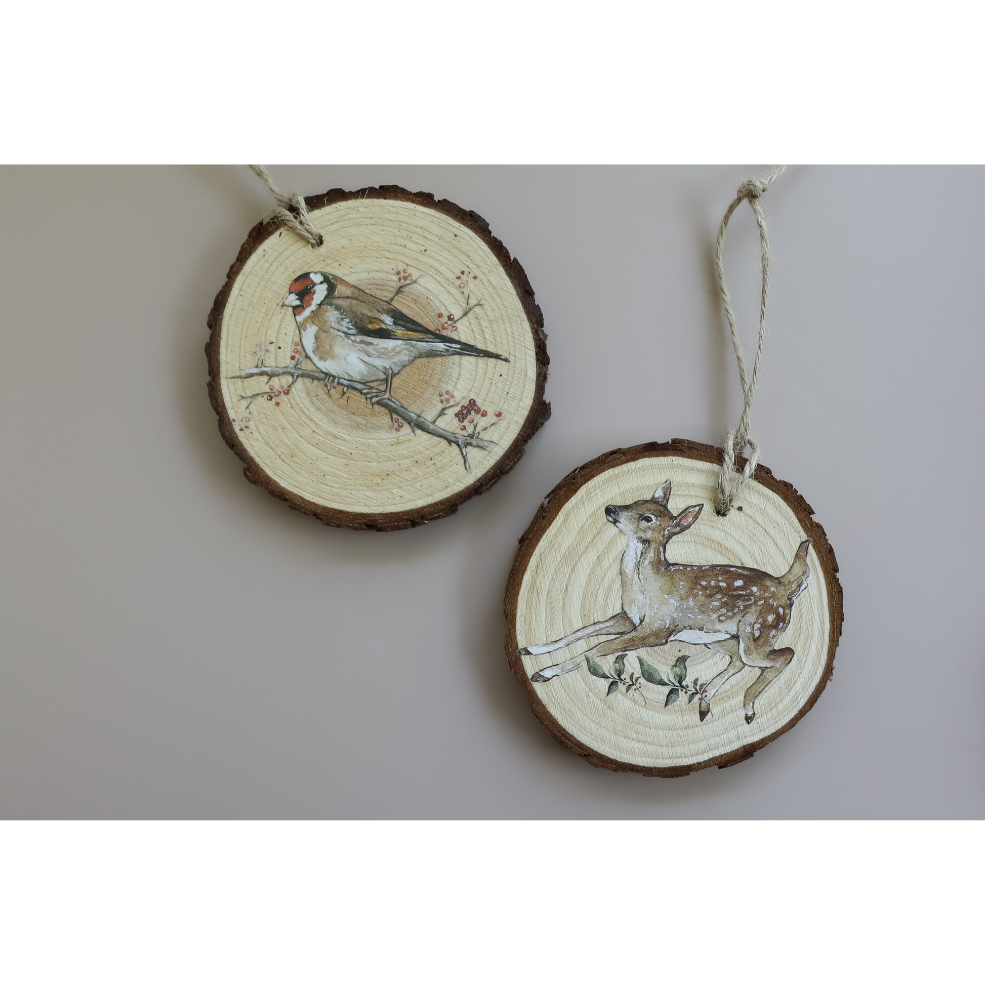 Two round wooden slice ornaments hanging on a light gray wall. One ornament features a goldfinch perched on a branch, and the other features a brown-and-white deer.
