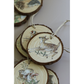 A cluster of round natural wood slice ornaments with bark edges, each decorated with woodland animal IOD transfer images such as deer, a bird, and mushrooms, hanging from twine on a light neutral background.