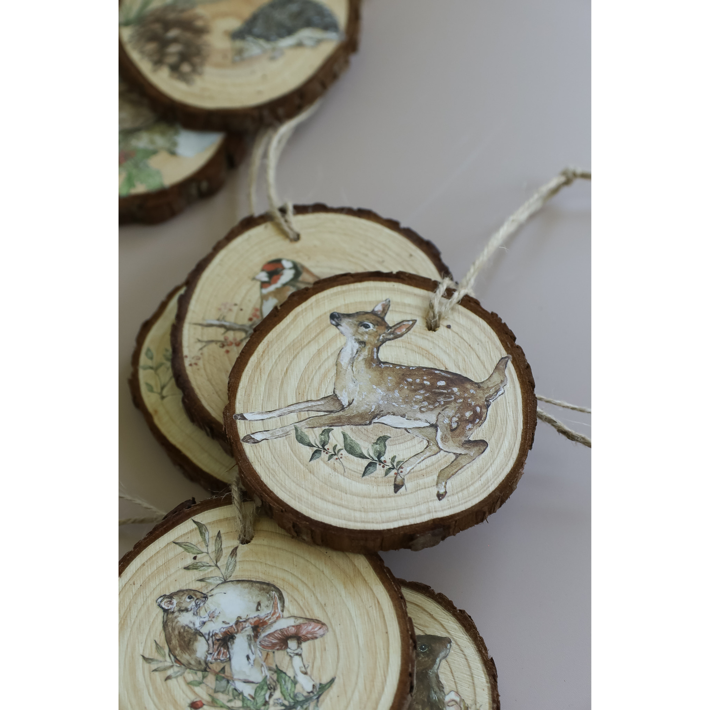 A cluster of round natural wood slice ornaments with bark edges, each decorated with woodland animal IOD transfer images such as deer, a bird, and mushrooms, hanging from twine on a light neutral background.