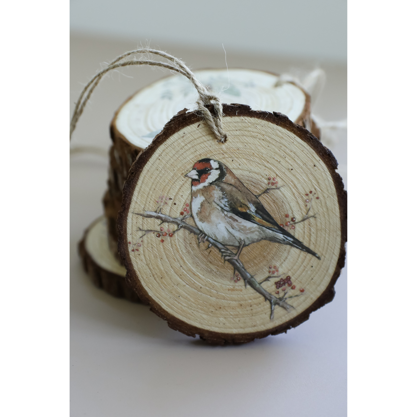 A round wooden slice ornament featuring an IOD transfer image of a bird on a branch, hanging from a twine loop, with other similar ornaments stacked behind it on a light neutral background.