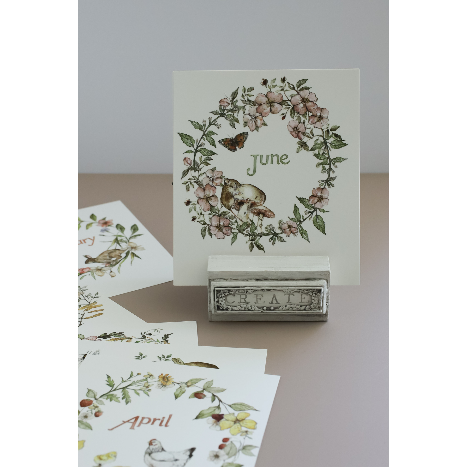 A botanical monthly calendar card for June displayed on a small stand, with other cards fanned out below showing IOD transfers of birds and flowers.