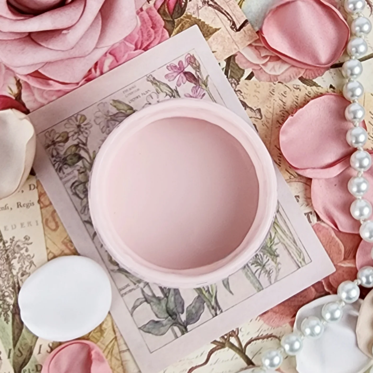 Image of an open jar of pale pink paint on a textured background with rose petals, pearls and a postcard.