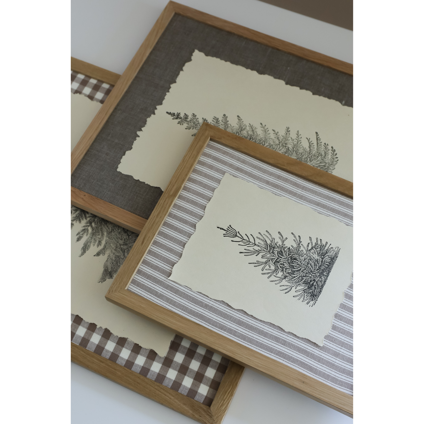 Three framed botanical art prints with light wood frames, arranged on a white surface. The prints feature black ink stamp impressions of greenery on cream paper, layered over striped and gingham fabric backdrops.