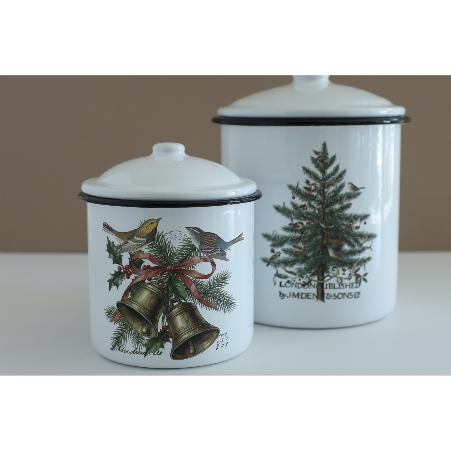 Two white enamel canisters with lids, decorated with IOD transfers. The smaller canister in the foreground features gold bells, holly leaves, pine branches, and birds tied with a red ribbon.