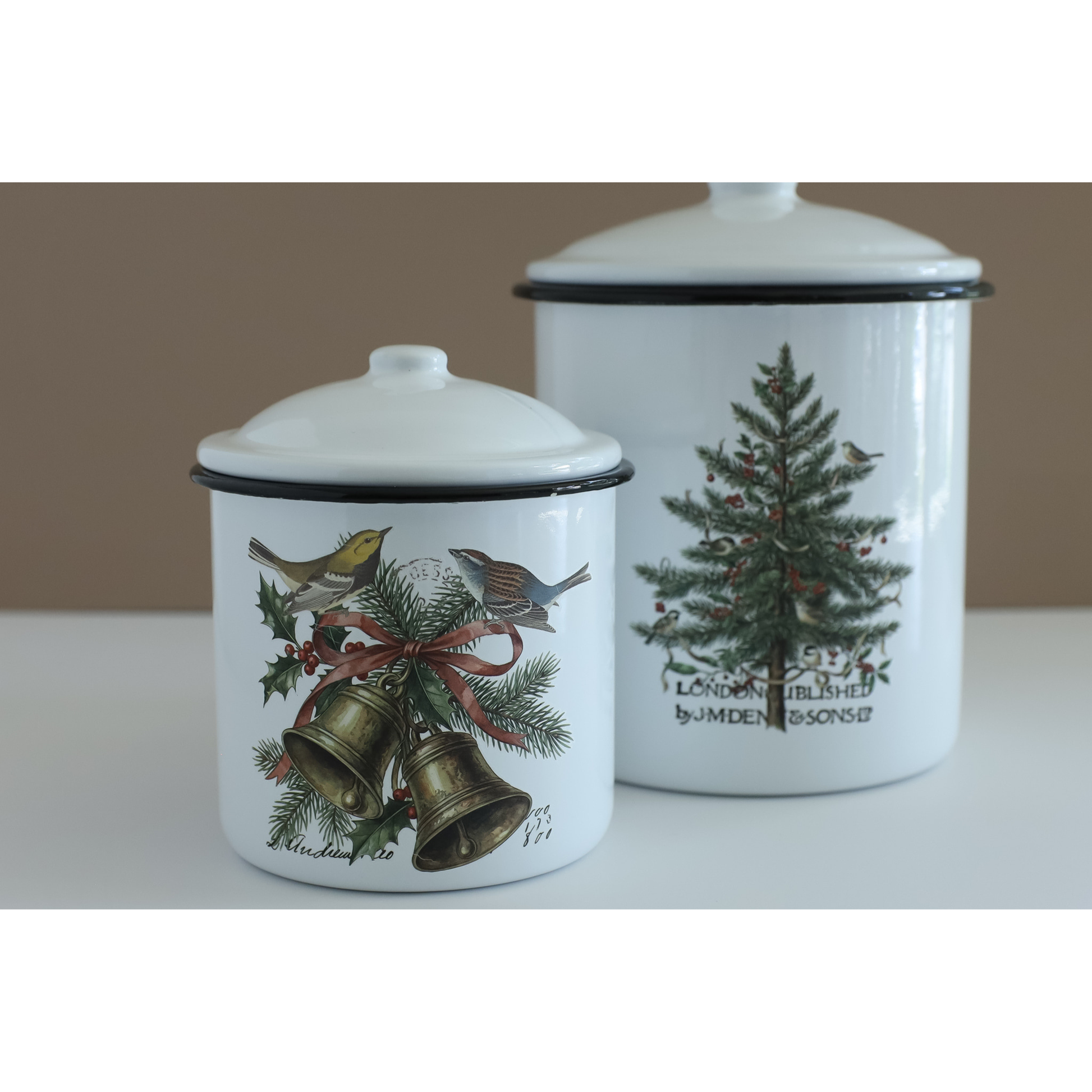 Two white enamel canisters with lids, decorated with IOD transfers. The smaller canister in the foreground features gold bells, holly leaves, pine branches, and birds tied with a red ribbon.