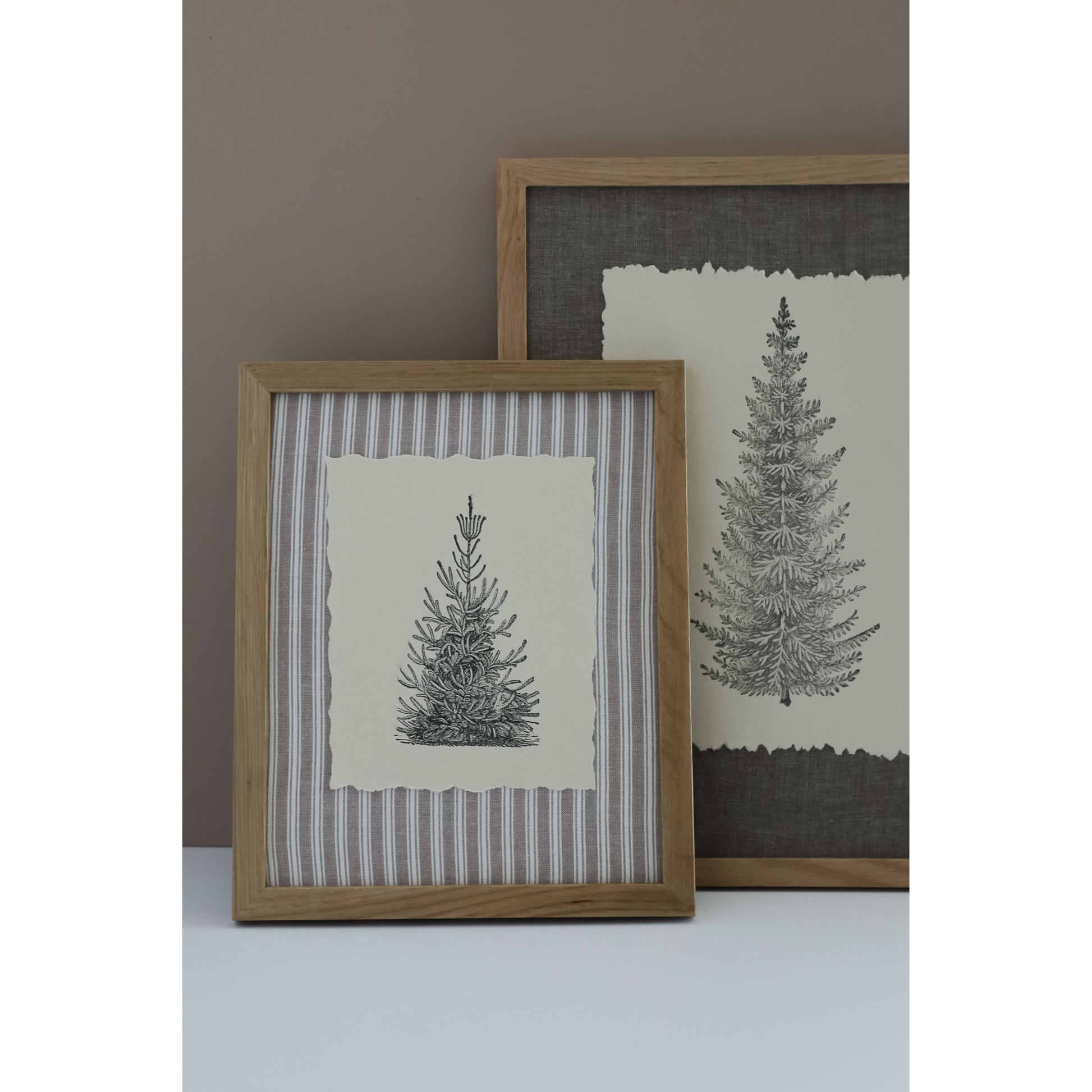 Two framed botanical art prints of pine trees, styled on a white surface against a taupe wall. The front print features a striped linen background with a central cream paper insert showing a black ink stamped image, enclosed in a light oak frame.