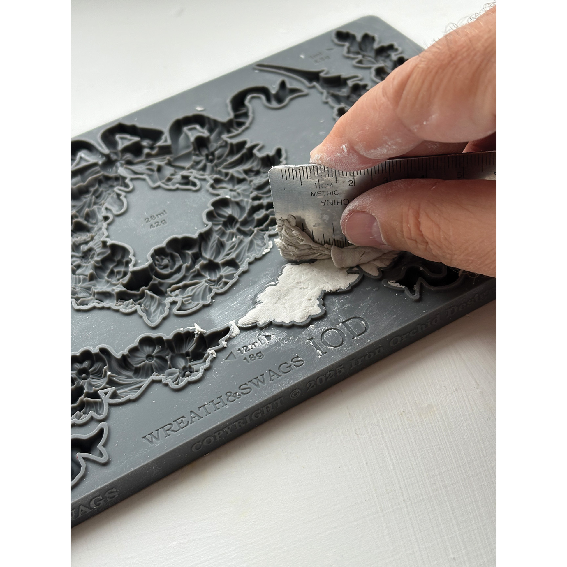 IOD Wreaths & Swags mold-product example. Hand using a tool to cut white material against a decorative gray mold.