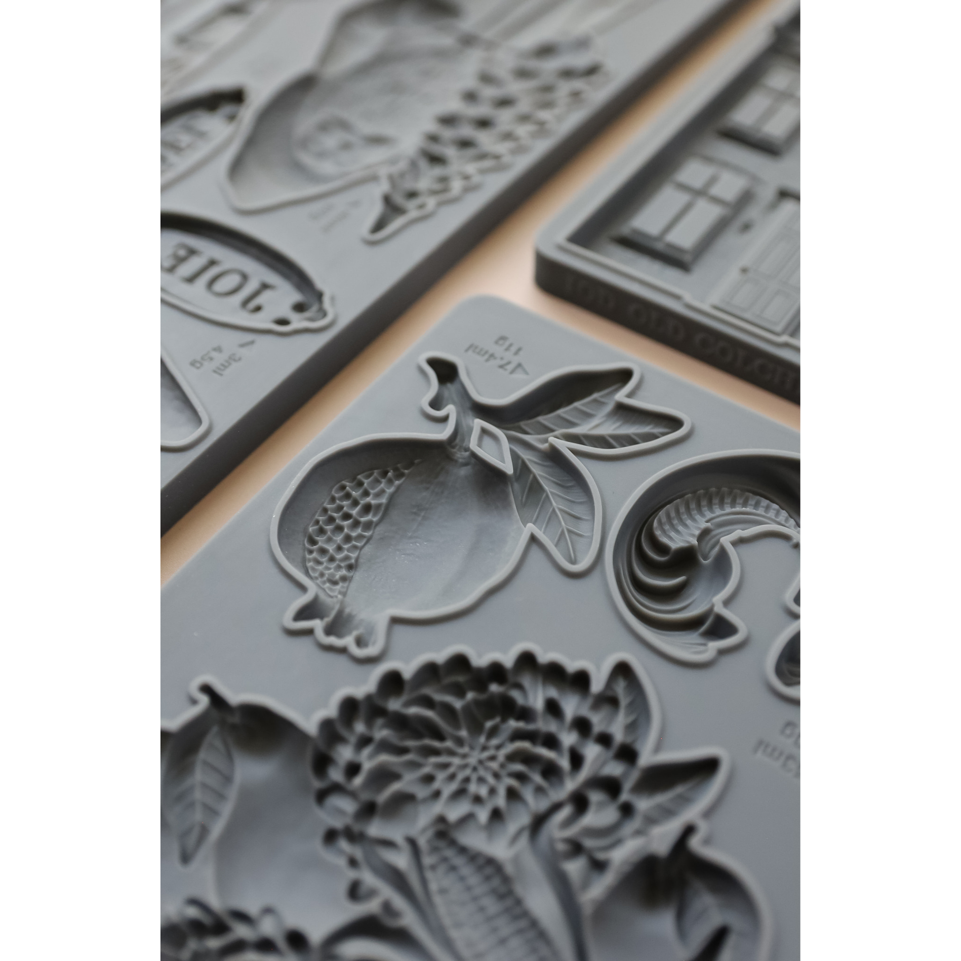 Gray silicone molds with embossed fruit and floral designs, arranged on a light beige surface.