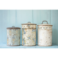 Three vintage-style metal canisters with floral patterns against a light blue wooden background